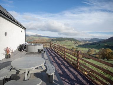 A wooden deck with a round picnic table and a hot tub overlooking green hills and a valley at Tyn Llwyn Barn in Glyndyfrdwy near Llangollen