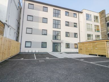 A parking lot with numbered spaces in front of a four-story apartment building with large windows and a wooden storage shed at Onshore 7 in Newquay