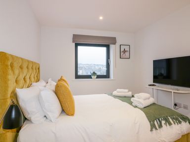 A bedroom with a yellow headboard bed white pillows and green blanket with towels on it a window a framed picture on the wall and a television on a white table at Onshore 7 in Newquay