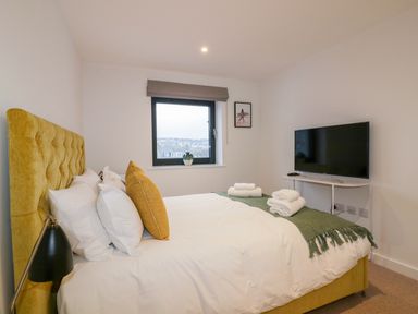 A bedroom with a yellow upholstered bed white bedding a green throw and a TV on a stand at Onshore 7 in Newquay