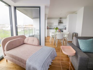 A living room with a pink chaise lounge and a gray sofa with a blue pillow near a dining table and white kitchen at Onshore 7 in Newquay