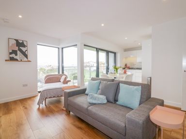 A living room with a gray sofa and pink side table and a small dining area and kitchen at Onshore 7 in Newquay