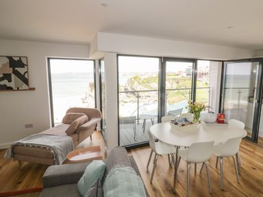 A dining area with a white table and six chairs next to large windows overlooking a beach and cliffside at Onshore 7 in Newquay