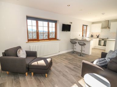 A living room with two grey armchairs and an ottoman near a window and an open kitchen with a breakfast bar and stools at Afon Rhos Bach in Llanrug