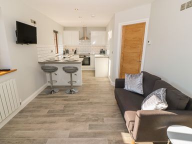A kitchen with a breakfast bar and two stools next to a living area with a sofa and cushions at Afon Rhos Bach in Llanrug