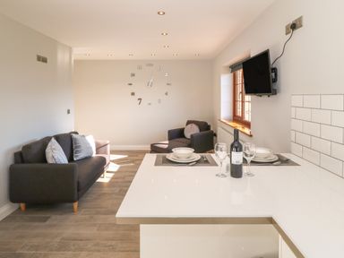 A living area with dark sofas, a wall clock, a mounted TV, a window, and a white counter with dining settings and a bottle of wine at Afon Rhos Bach in Llanrug