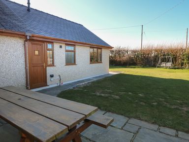 An outdoor area with a wooden picnic table, a house with brown door and windows, a grassy lawn, and a swing in the garden at Afon Rhos Bach in Llanrug