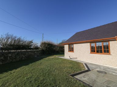 A side yard with grass and a stone wall next to a building with two windows at Afon Rhos Bach in Llanrug