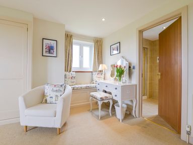 A room with a white armchair a built-in window seat with cushions a white vanity table with a mirror and stool a lamp and flowers and an open door to a bathroom at Duffield in St Ann's Chapel
