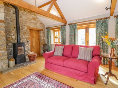 A living room with a red sofa two striped cushions a wood stove a stone wall wooden beams floral curtains and a side table with flowers at Middle Farm Annex in Long Bredy