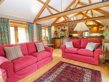 A living room with two red sofas and a patterned rug with visible wooden beams and a kitchen in the background at Middle Farm Annex in Long Bredy