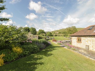 A garden with a lawn pathway flowering bushes a stone wall wooden seating and a stone building at Middle Farm Annex in Long Bredy