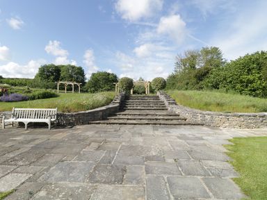 An outdoor stone stairway leading up to wooden pergolas with greenery and a wooden bench on the left at Middle Farm Annex in Long Bredy