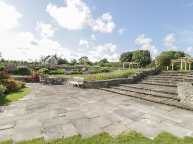 An outdoor patio area with stone steps wooden benches and a dining set surrounded by greenery and flowers at Middle Farm Annex in Long Bredy