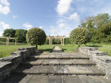 Stone steps leading to a pathway with two trimmed round trees and wooden pergolas in a grassy area at Middle Farm Annex in Long Bredy