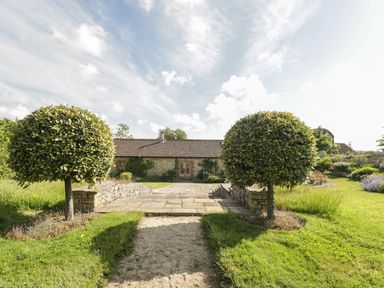A garden with two round pruned trees and stone path leading to a stone building with tiled roof at Middle Farm Annex in Long Bredy