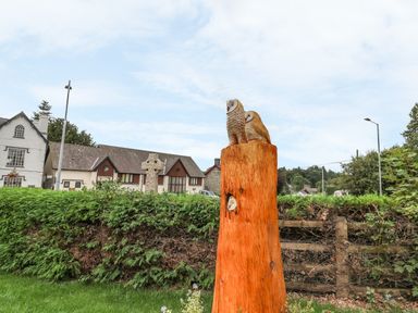 An outdoor scene showing a wooden sculpture of two owls perched on a tree stump near a hedge and residential buildings at New Peny in Llandrindod Wells