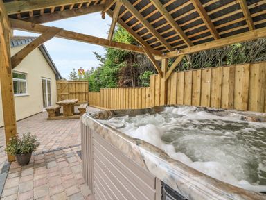 A hot tub under a wooden pergola next to a wooden picnic table on a paved patio at New Peny in Llandrindod Wells