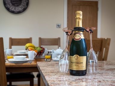 A kitchen countertop with a bottle of Taittinger champagne and two glasses hanging on it with a dining table set with dishes and fruit bowl in the background at New Peny in Llandrindod Wells