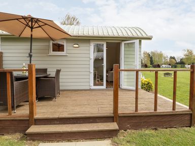 A wooden deck with a table umbrella and chairs outside a small building with glass doors at Bramble in Banham