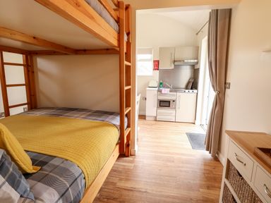 A small kitchen area with white cabinets and a microwave visible through an open doorway from a room with wooden bunk beds with plaid bedding at Bramble in Banham