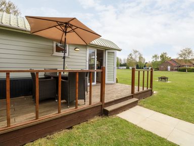 A wooden deck with outdoor table and umbrella next to a light green building with open doors and views of grass and trees at Bramble in Banham