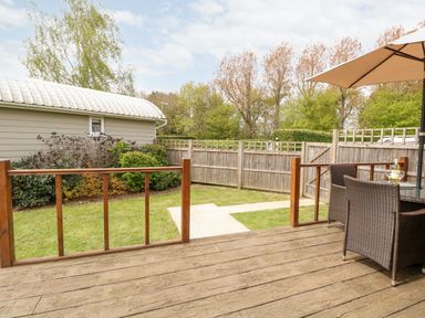 A wooden deck with table umbrella and chairs overlooking a fenced garden with bushes and a shed at Bramble in Banham