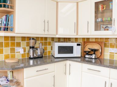 A kitchen corner with cream cabinets yellow tiled backsplash a microwave a food processor and a vintage scale at 1 Orchard Cottages in Salcombe