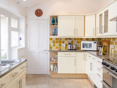 A kitchen with cream cabinets yellow tiled backsplash a microwave a food processor and a clock above a white door at 1 Orchard Cottages in Salcombe