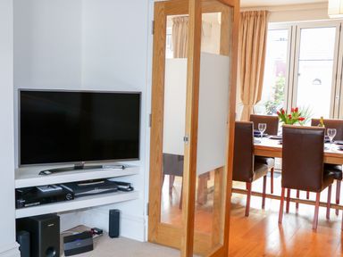 A corner with a wall-mounted television and electronic devices next to a wooden dining table with chairs in a room at 1 Orchard Cottages in Salcombe