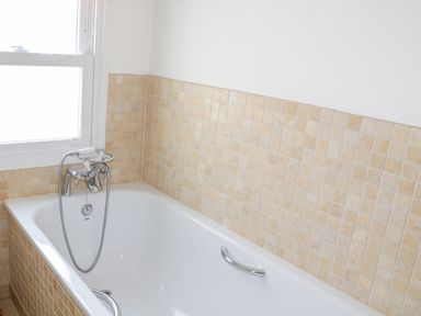 A bathroom with a white bathtub and beige tiled walls at 1 Orchard Cottages in Salcombe