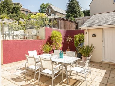 An outdoor patio with a white table and six chairs surrounded by potted plants and a red wall at 1 Orchard Cottages in Salcombe