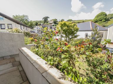 A garden with flowers and shrubs next to a paved patio area with houses and trees in the background at 1 Orchard Cottages in Salcombe
