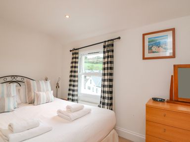 A bedroom with a bed with folded towels on it a window with checkered curtains and a wooden dresser with a mirror at 1 Orchard Cottages in Salcombe