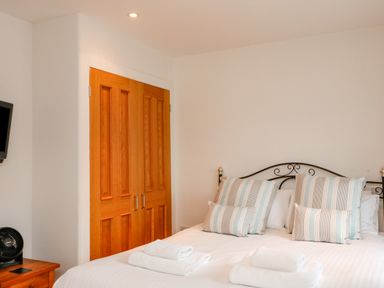 A bedroom with a double bed with striped pillows folded towels wooden wardrobe and a small fan at 1 Orchard Cottages Salcombe