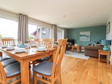 A dining table with wooden chairs and tableware next to a living room with sofas and a coffee table at Lodge Sixty Six in St Columb Major
