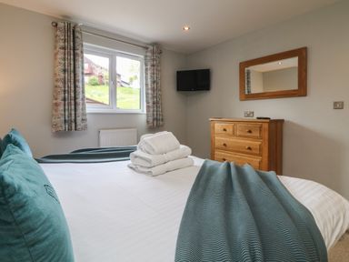 A bedroom with a bed with teal pillows and blanket folded towels on the bed a wooden dresser a wall-mounted tv and a window with curtains at Lodge Sixty Six in St Columb Major