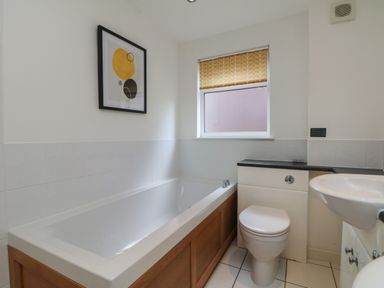 A bathroom with a bathtub, toilet, sink, window with a patterned blind, and a framed abstract artwork on the wall at Lodge Sixty Six in St Columb Major