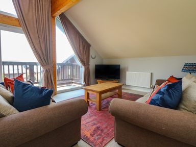 A living room with two brown sofas blue and red cushions a wooden coffee table a flat screen TV and large windows with curtains at Lodge Sixty Six in St Columb Major