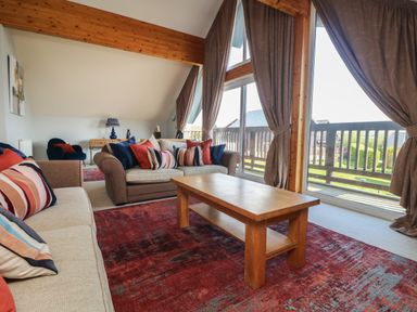 A living room with sofas and a wooden table on a red rug with large windows and curtains at Lodge Sixty Six in St Columb Major