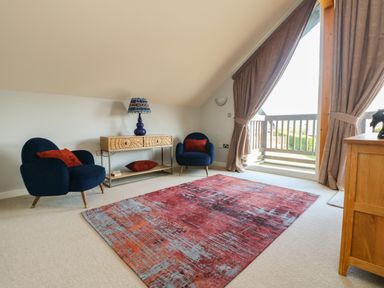 A room with two blue chairs with red pillows a wooden console table with a lamp a red and blue rug and large curtains at Lodge Sixty Six in St Columb Major