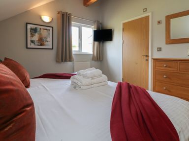 A bedroom with a bed with red pillows and throw a window with curtains a wall mounted TV a wooden dresser and a door at Lodge Sixty Six in St Columb Major