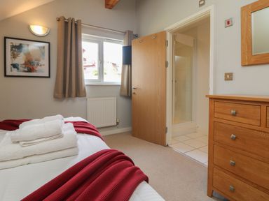 A bedroom with a bed with white and red bedding a wooden dresser a window with curtains and an open door to a bathroom at Lodge Sixty Six in St Columb Major