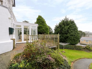 A conservatory attached to a house with a wooden deck garden shrubs and trees at Preswylfa Apartment in Caernarfon