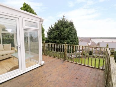 A wooden deck with railing overlooking a garden and houses with water in the background at Preswylfa Apartment in Caernarfon