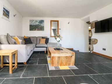 A living room with a grey sectional sofa wooden coffee table with flowers a wall-mounted television and stone flooring at Miner's Rest in Troon near Camborne