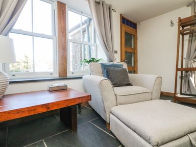 A sitting area with an armchair and ottoman near a wooden table and large window at Miner's Rest in Troon near Camborne