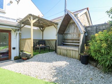 A backyard with a wooden gazebo, plastic chairs, a table with a tea set, a wooden bench with a roof, gravel ground and plants at Miner's Rest in Troon near Camborne