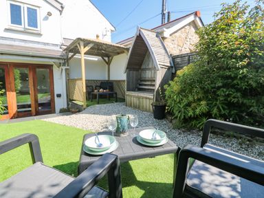 An outdoor patio area with artificial grass a small table set for two wooden seating under a canopy and a wooden shelter at Miner's Rest in Troon near Camborne