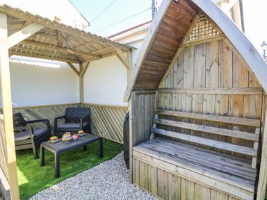 An outdoor seating area with two black chairs and a black table with a colorful tea set under a wooden pergola next to a wooden bench with a pitched roof at Miner's Rest in Troon near Camborne